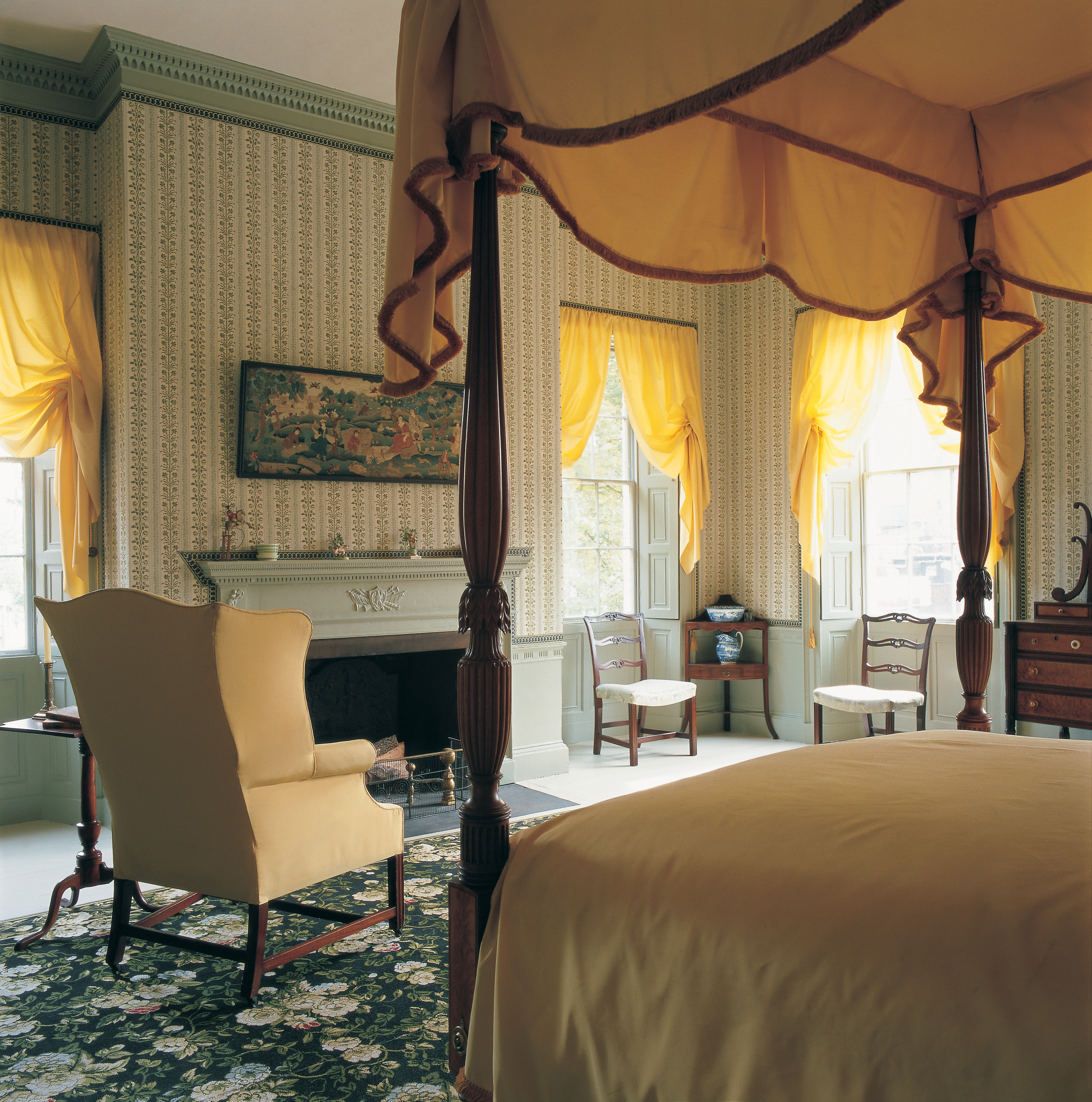 Room with patterned wallpaper, yellow curtains, and various pieces of wooden furniture. 