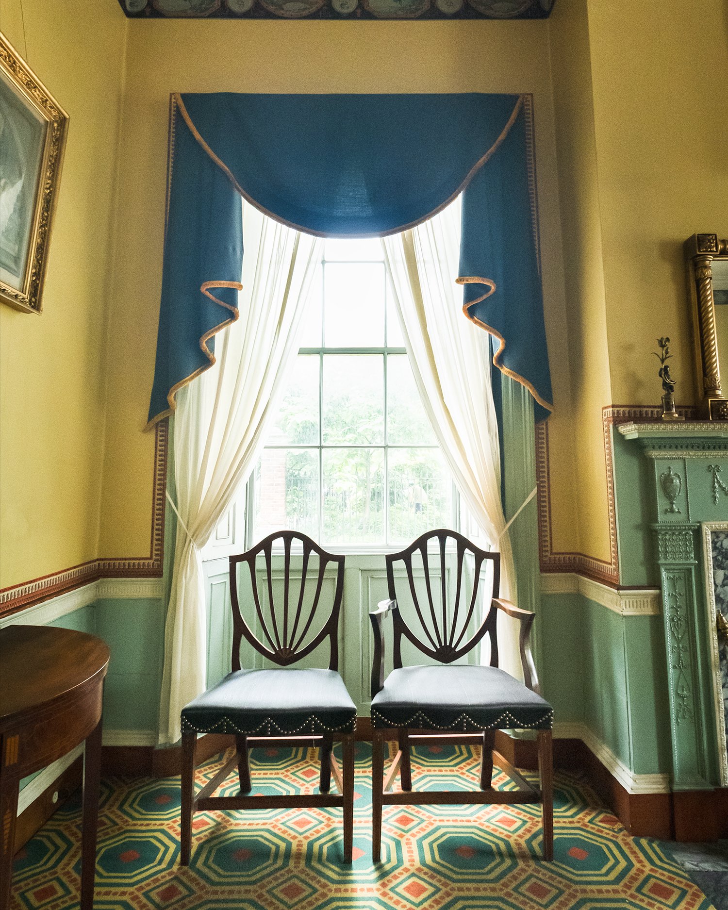 Two dark-wood chairs with a shield-shaped back, the one on the right has arms, placed in front of a window and framed by sheer curtains. The surrounding walls are a grey-green painted color on the bottom with a yellow wallpaper with a red border at the to